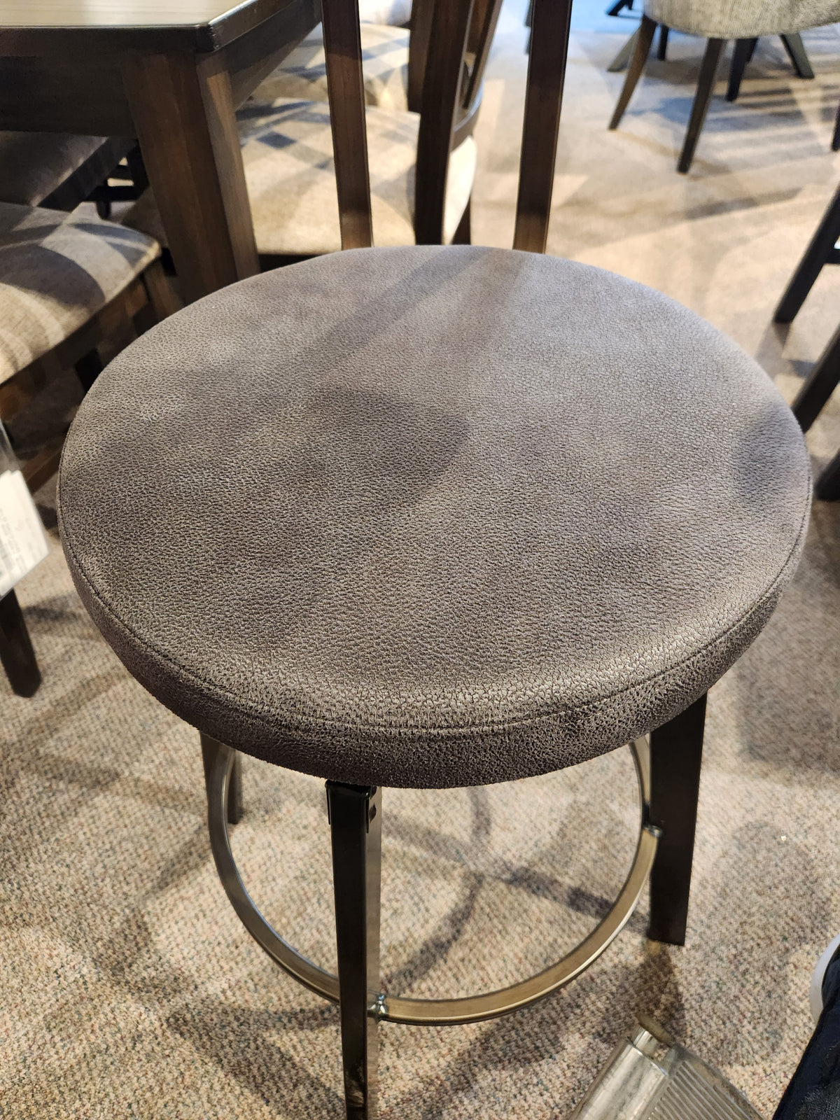 The Amisco Architect Counter Height Stool has a round padded seat, dark metal legs, and is shown on carpet in-store.
