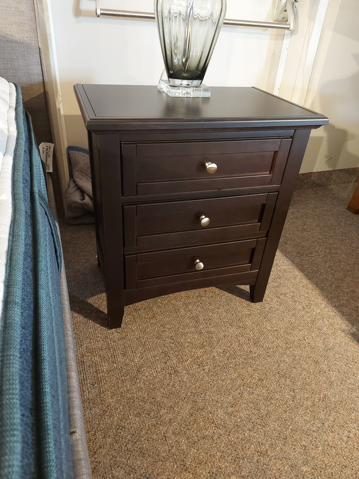 The Vaughan Bassett BB27 Merlot Nightstand features three drawers with silver knobs and sits on a carpeted floor by the bed.
