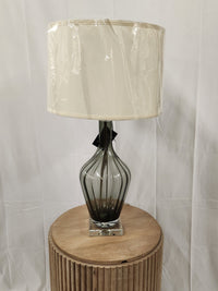 The Pacific Coast 790A4 Nile Glass Table Lamp with a vase silhouette in smoky gray glass sits on a round wood table.