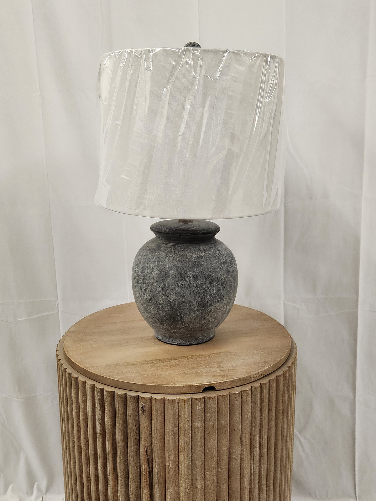 The 862D0 Anza Table Lamp by Pacific Coast, in gray ceramic with a drum shade, rests on a round wooden table.