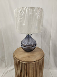 The Ashley L430784 Lemmitt Table Lamp features a transparent blue glass base and a white shade on a wooden pedestal.
