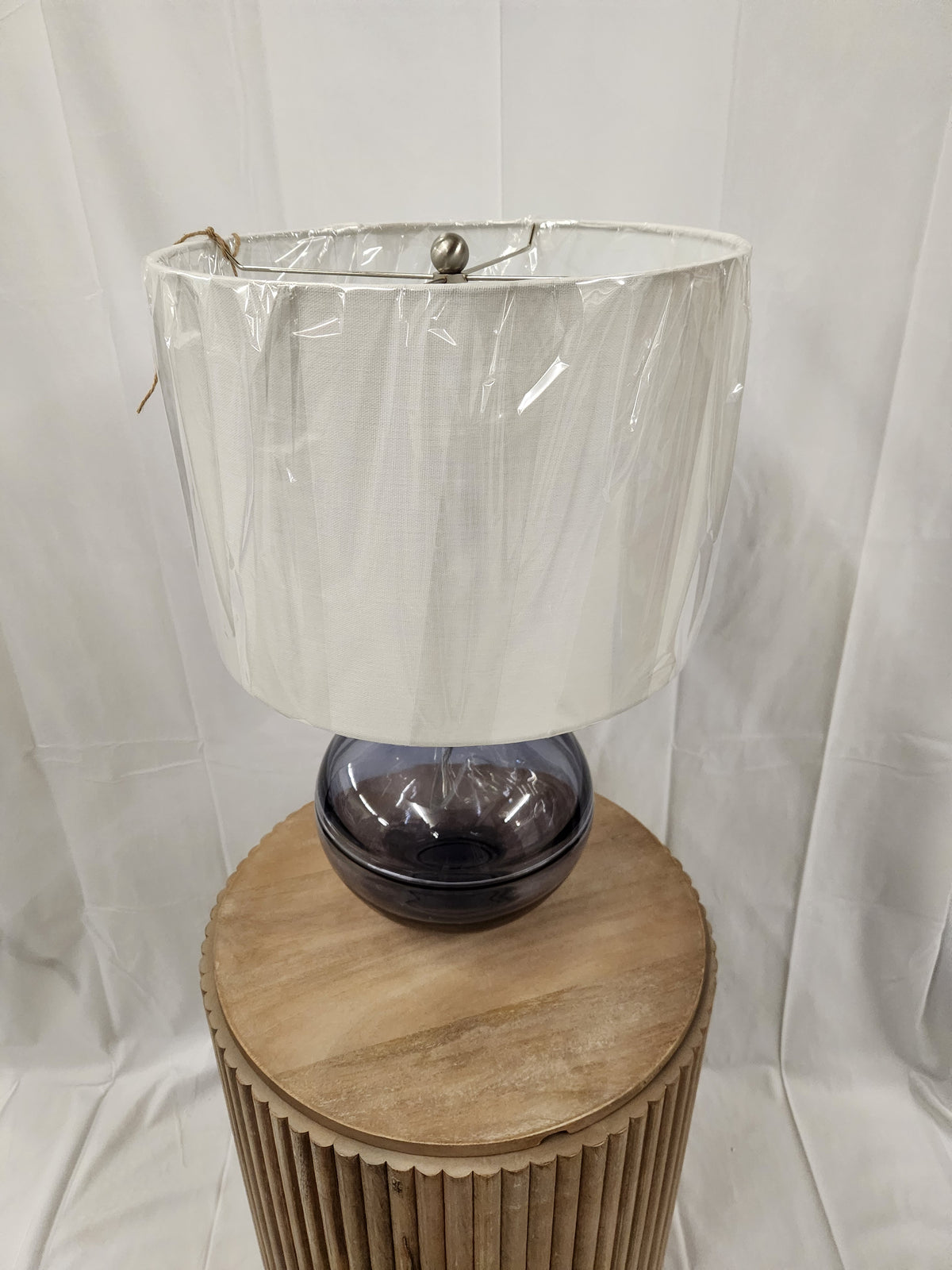 The Ashley L430784 Lemmitt Table Lamp with a transparent blue finish and white shade sits on a round wooden stool.