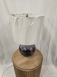 The Ashley L430784 Lemmitt Table Lamp with a transparent blue finish and white shade sits on a round wooden stool.