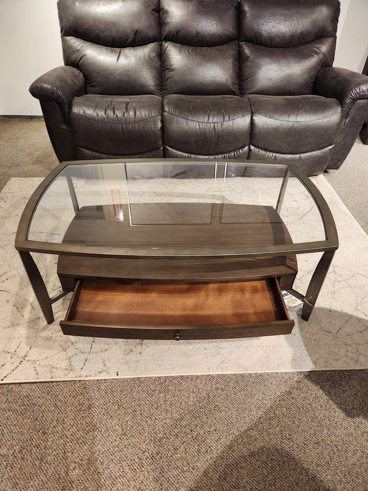 The Magnussen T5692 Ardis Coffee Table, with oak veneer and glass top, sits in front of a dark sofa.