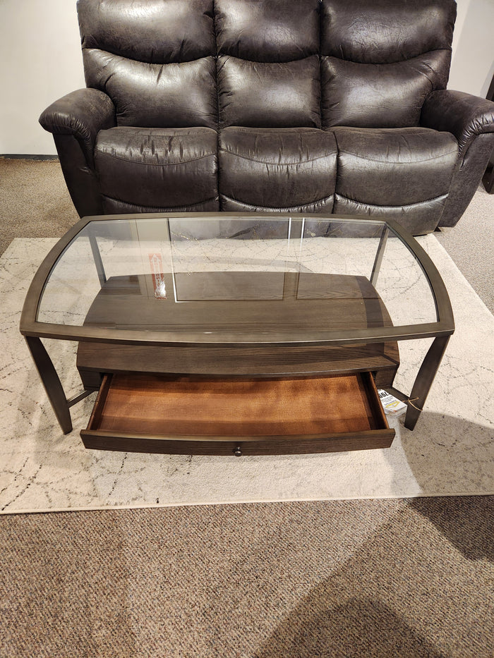 The Magnussen T5692 Ardis Coffee Table, with oak veneer and glass top, sits in front of a dark sofa.