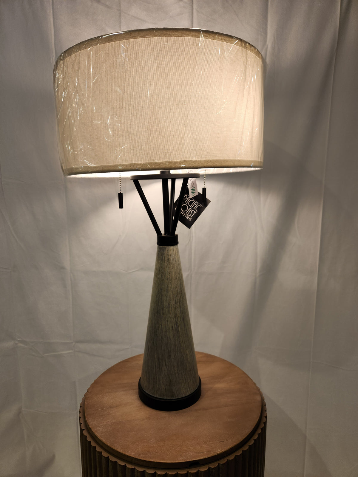 The 71N94 Oakland Table Lamp by Pacific Coast radiates industrial charm, featuring a cone-shaped base and a spacious beige fabric lampshade as it elegantly rests on a round wooden surface.