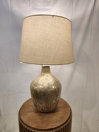 The Ashley Mari Table Lamp with a textured beige shade radiates vintage vibes on a round wooden table.