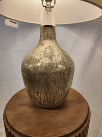 The Mari Table Lamp by Ashley, with a textured mercury glass base and beige shade, sits on a circular wooden table.
