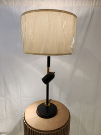 The Pacific Coast 96J17 Holmes Table Lamp in matte black with a beige shade is displayed on a round wooden table.