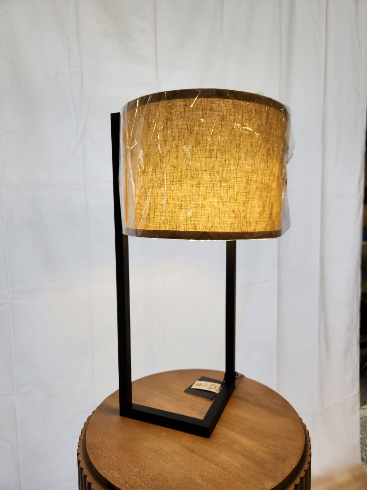 The Thorton Black Table Lamp by Pacific Coast features a sleek black metal frame and beige shade on a round wooden table.