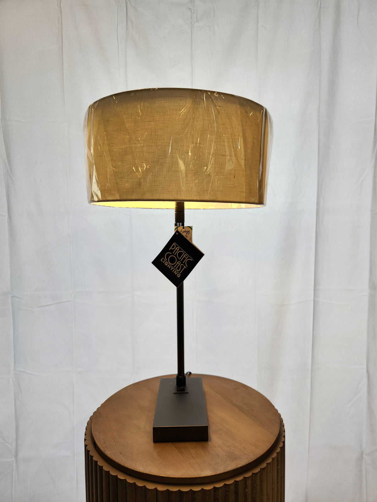 A Pacific Coast 74G04 HUGO Modern Desk Lamp with beige shade, black base sits on a wood round table; shade in plastic.