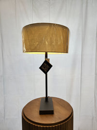 A Pacific Coast 74G04 HUGO Modern Desk Lamp with beige shade, black base sits on a wood round table; shade in plastic.