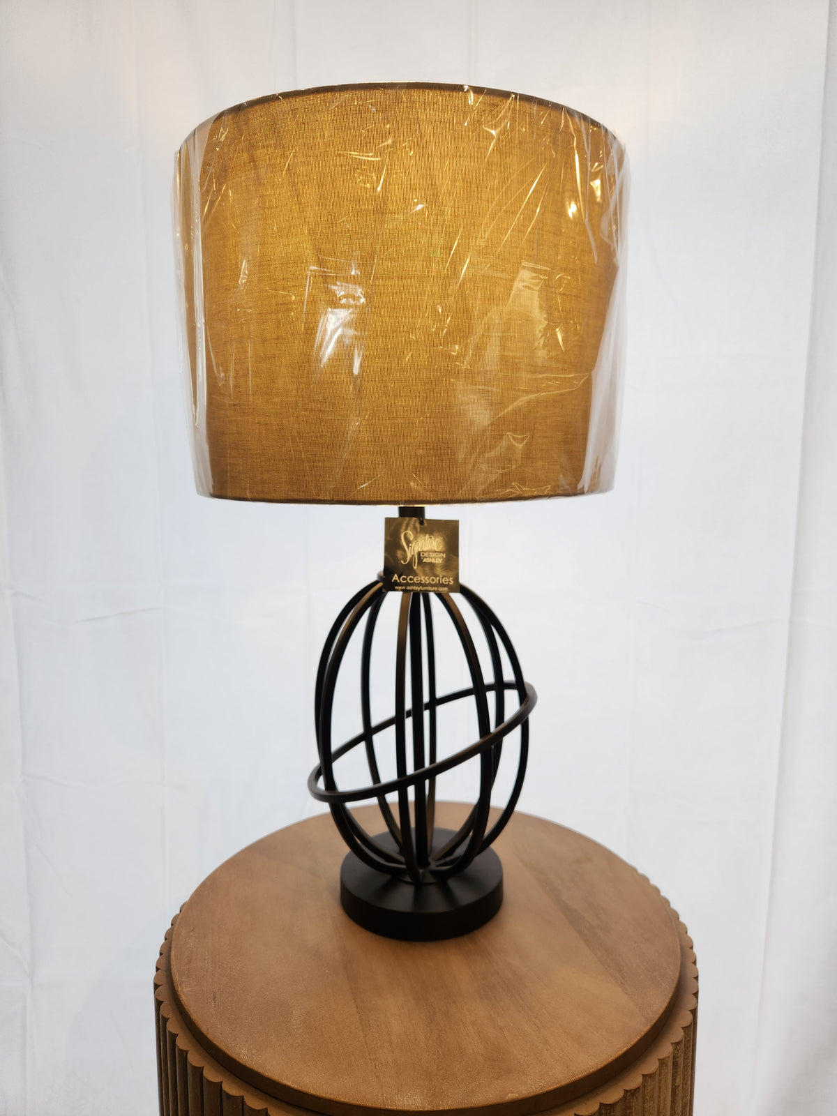 The Ashley L204164 Manasa Table Lamp with a round brown shade sits on a wooden circular side table.