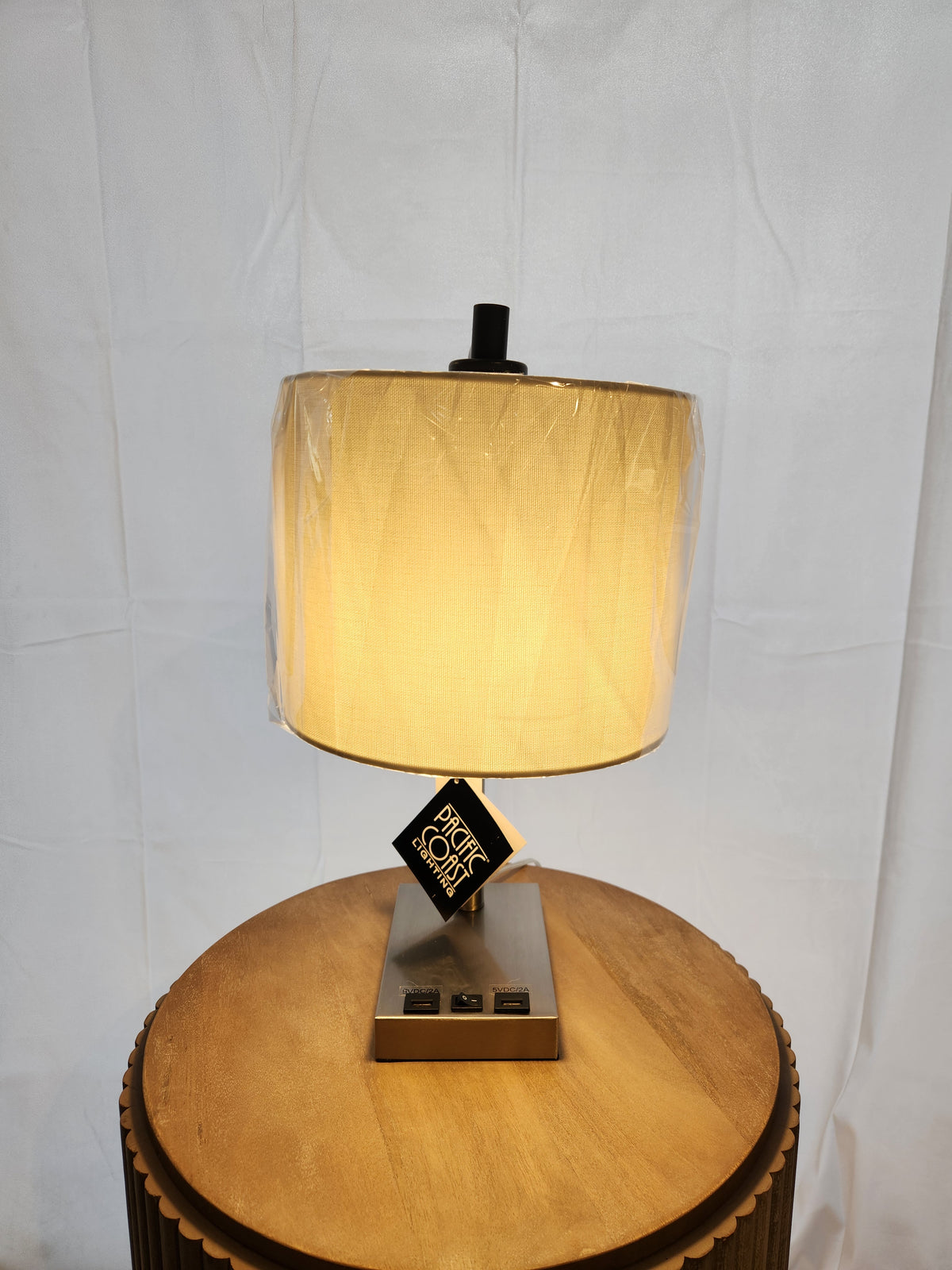 The Pacific Coast 902c0 Elton Table Lamp sits on a round wood side table, with a linen shade and USB ports on its base.