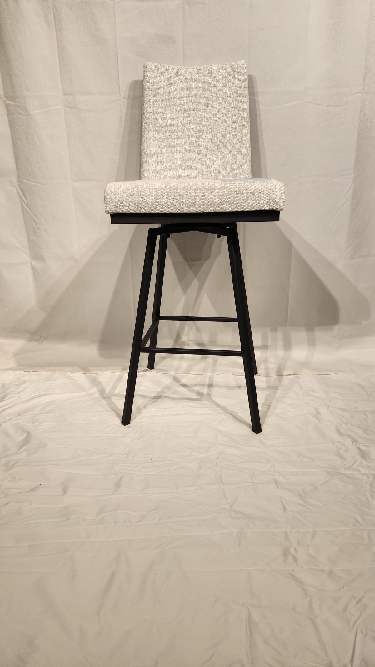 The Amisco Linea light gray swivel bar stool with a metal frame stands on a white fabric backdrop.