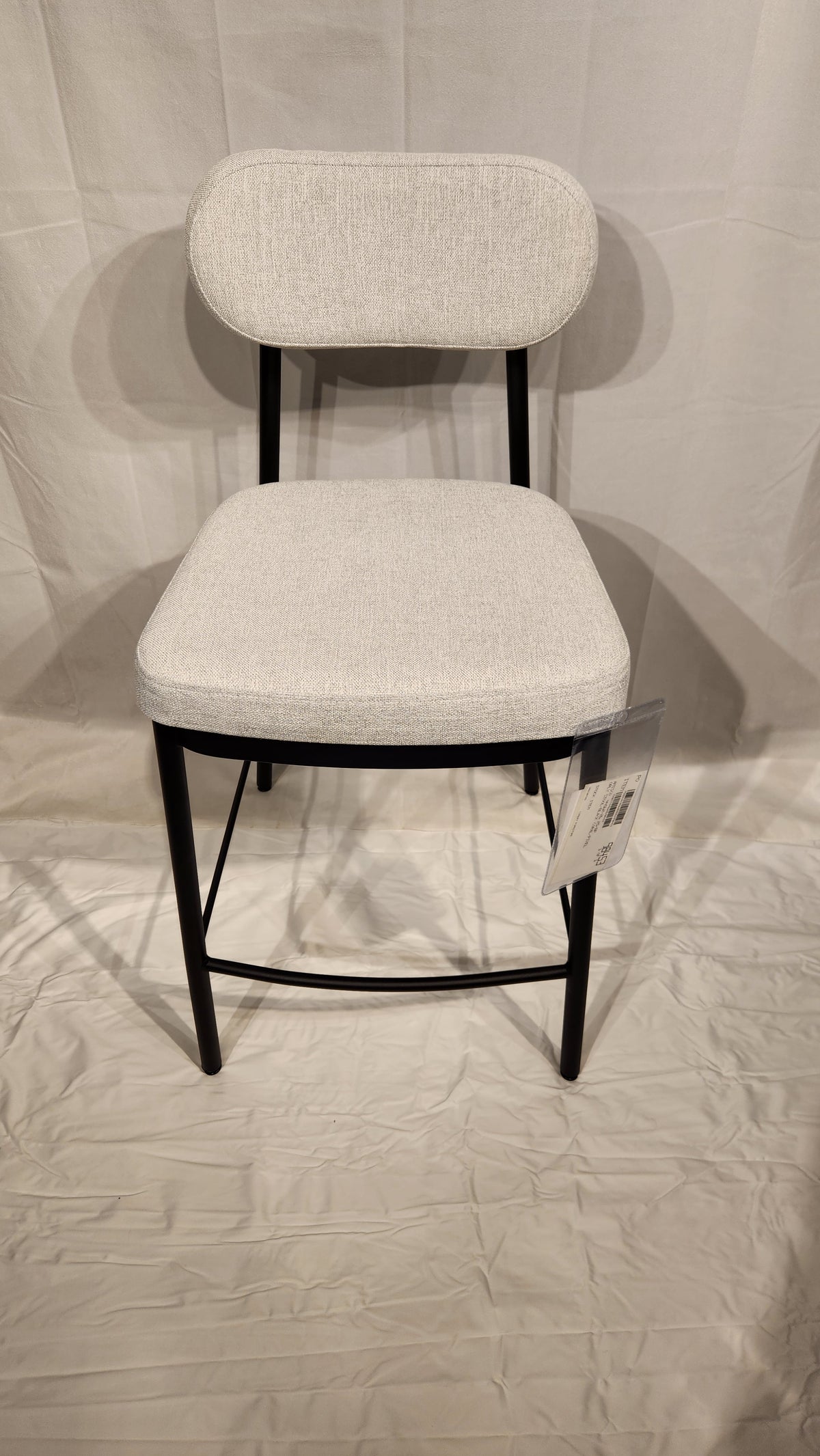The Amisco Orly Counter Height Bar Stool features a light gray cushioned seat, backrest, and black metal legs on white.