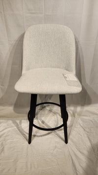 A Zahra Counter Height Swivel Bar Stool by Amisco, beige with black metal legs, sits on a white fabric backdrop with a tag.