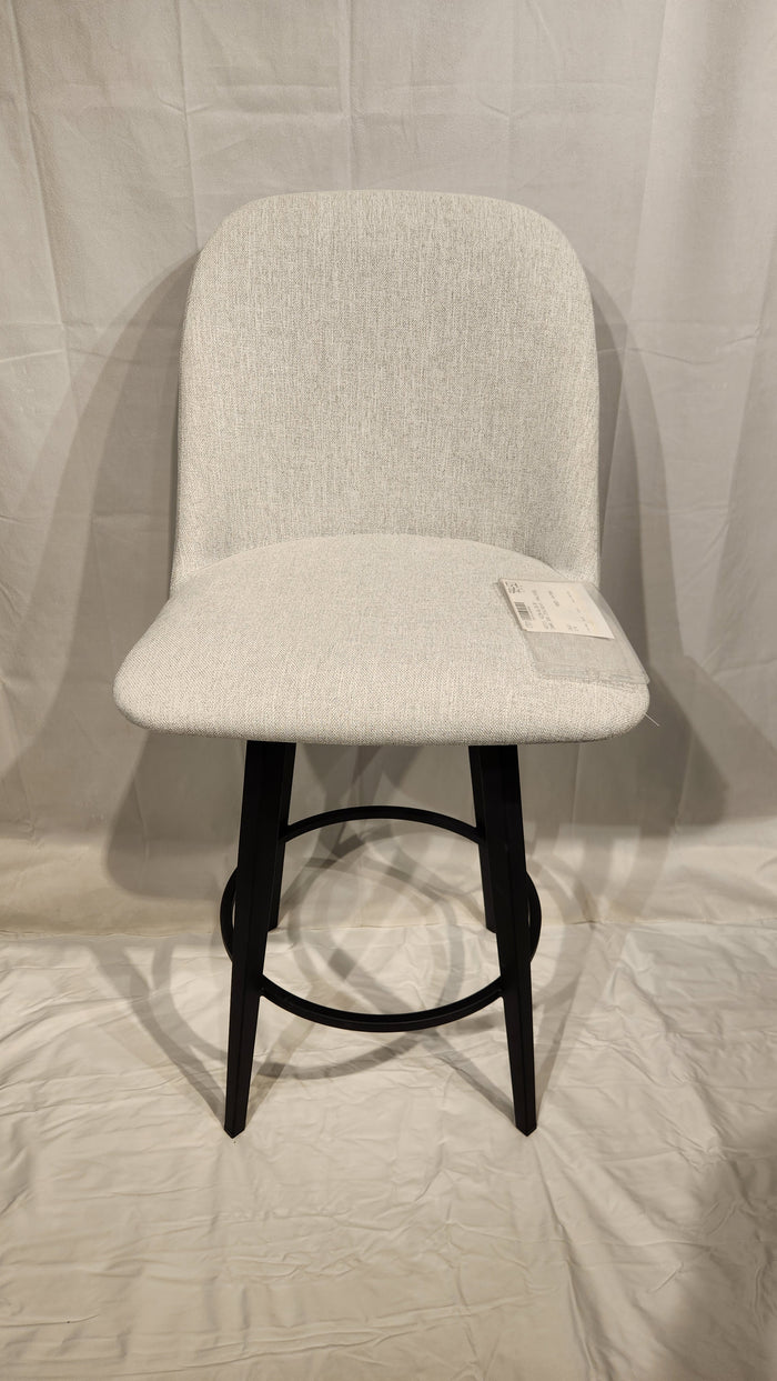 A Zahra Counter Height Swivel Bar Stool by Amisco, beige with black metal legs, sits on a white fabric backdrop with a tag.