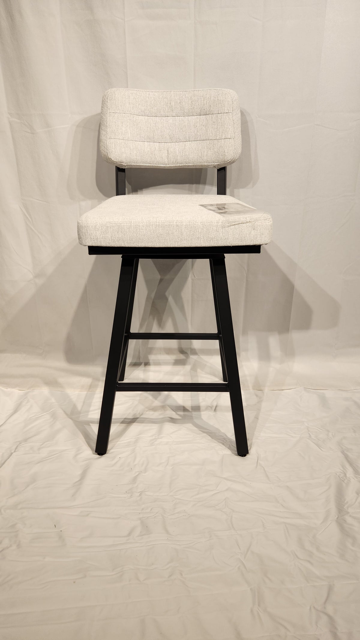 The Phoebe Bar Stool by Amisco, with a white cushion and black frame, is displayed on a white cloth backdrop.