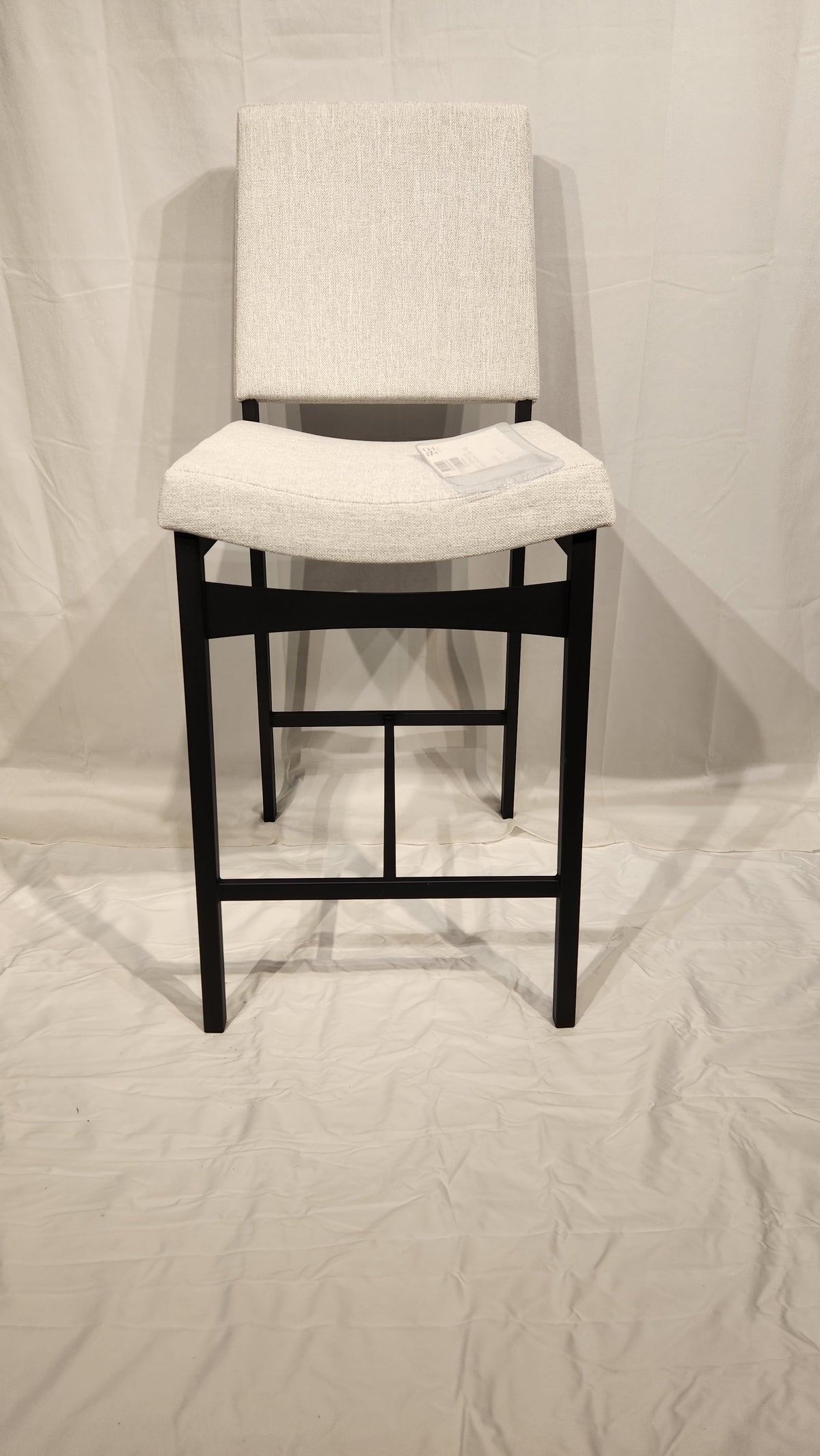The Amisco Kendra Counter Height Bar Stool in light gray upholstery with a metal frame stands on a white sheet-covered floor.