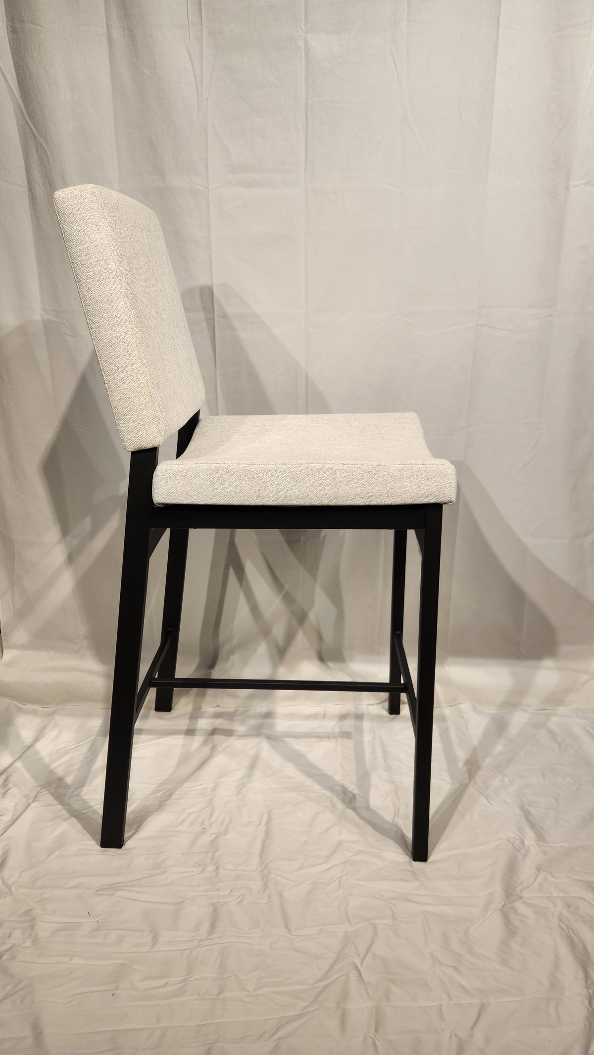 Side view of an Amisco Kendra Counter Height Bar Stool with a light beige seat and backrest, featuring black wooden legs.