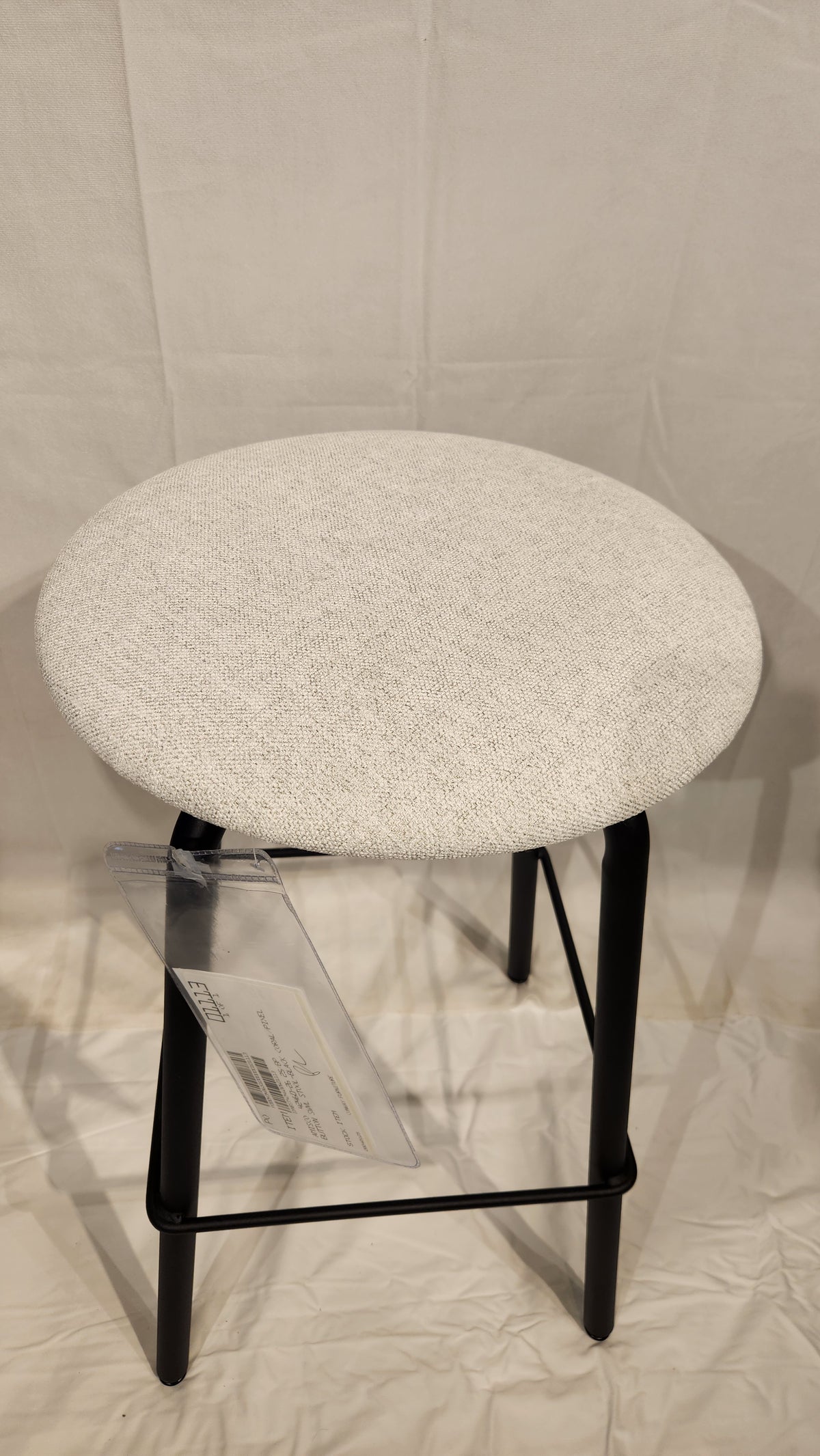 The Amisco Button Counter Height Swivel Stool features a round white seat and black metal legs on a light fabric background.