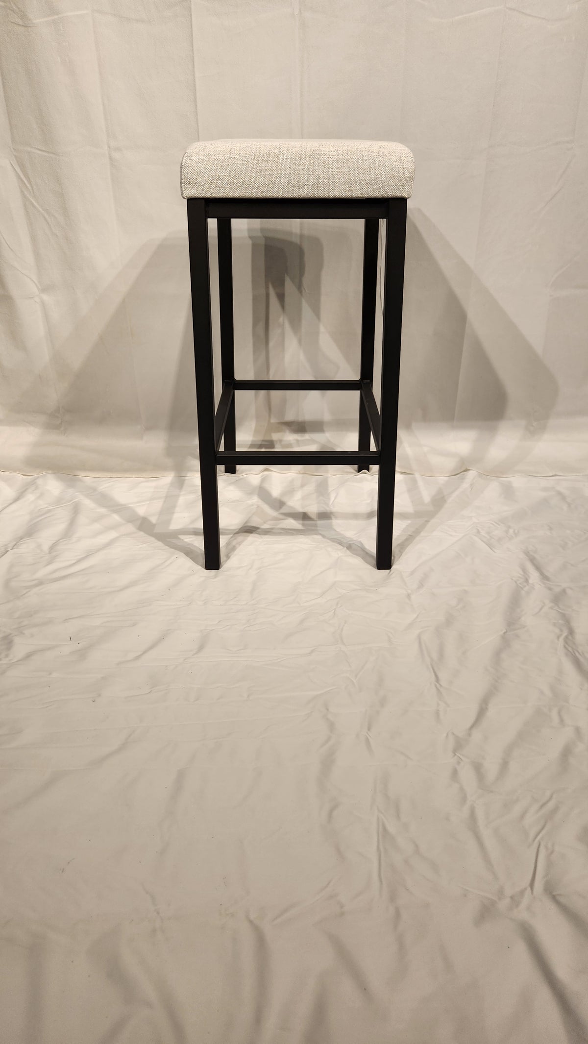 The Amisco Bradley Counter Height Bar Stool features a tall black metal frame and light upholstered seat on display.