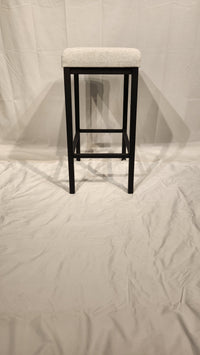 The Amisco Bradley Counter Height Bar Stool features a tall black metal frame and light upholstered seat on display.