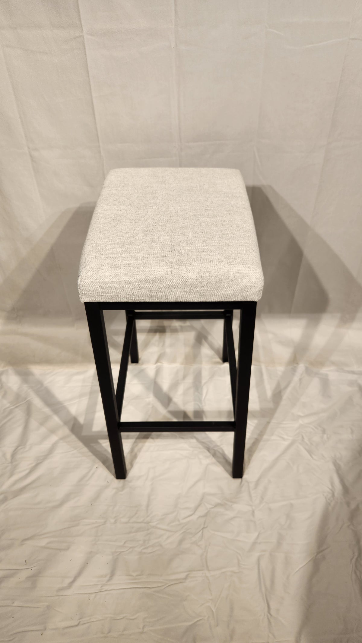 The Amisco Bradley Counter Height Bar Stool features square, light beige upholstery and black metal legs on white fabric.