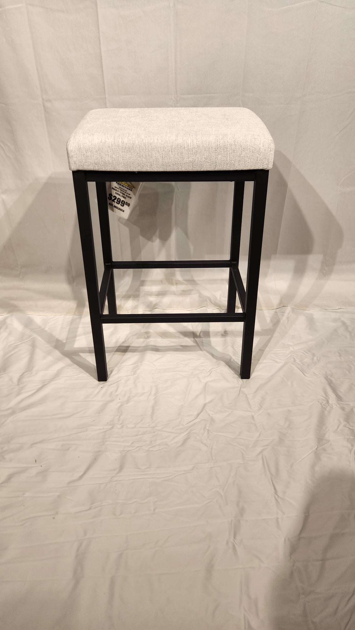 The Bradley Counter Height Bar Stool by Amisco features a light seat and black metal legs on a white fabric background.
