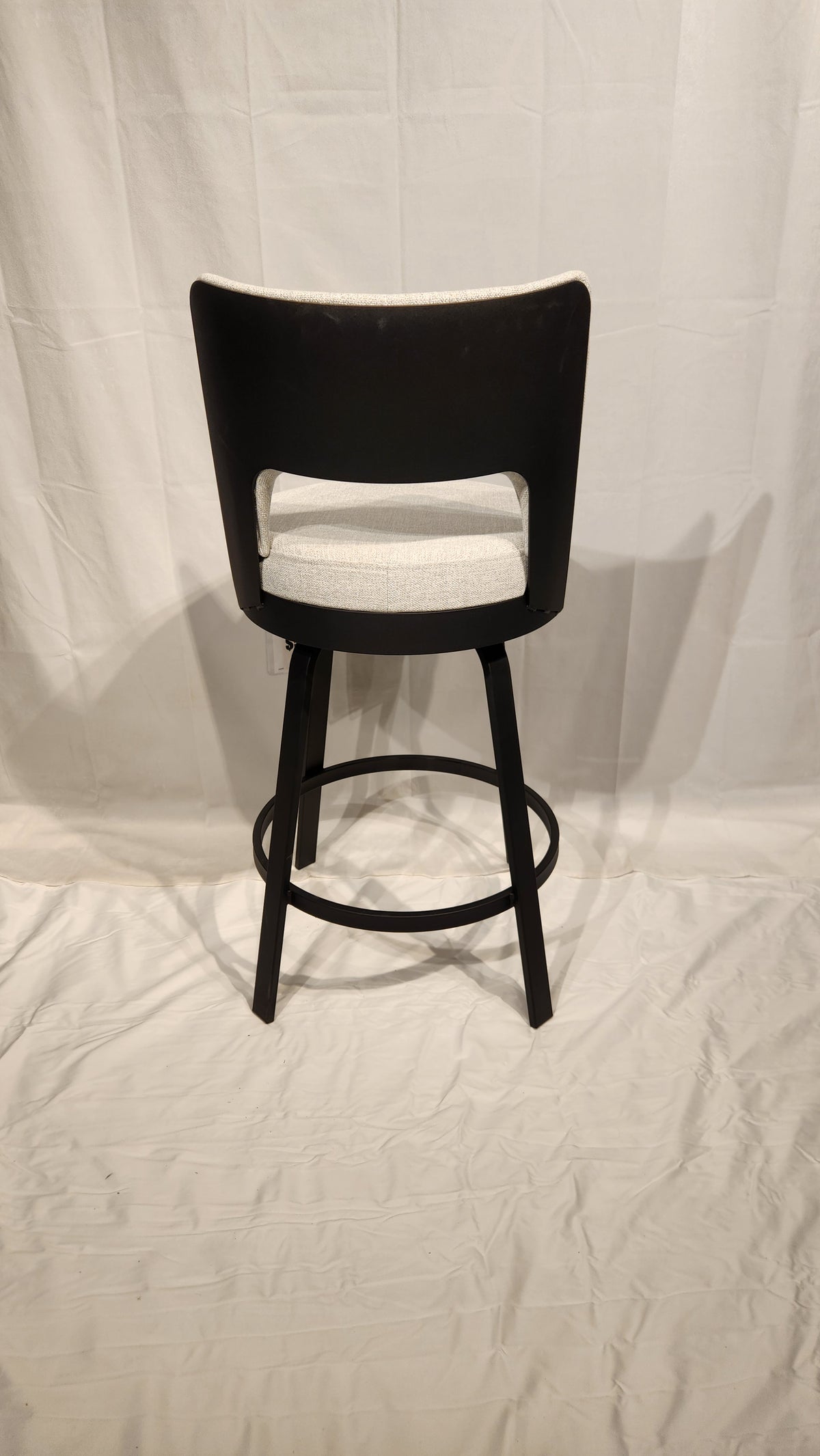 The Amisco Brock Counter Height Swivel Bar Stool, with a backrest, is shown from behind on a white fabric backdrop.