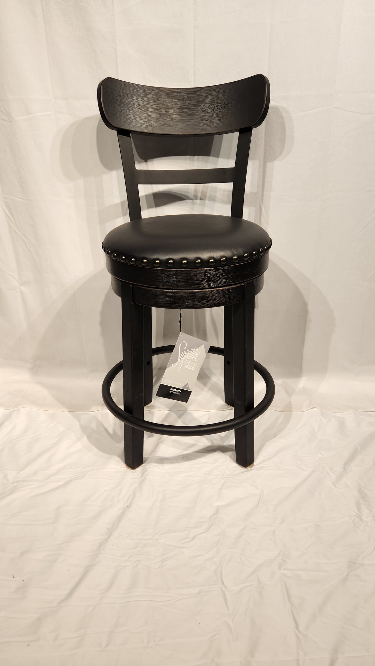 An Ashley Valebeck D546 black wood bar stool features a cushioned round seat, metal footrest, and nailhead trim.