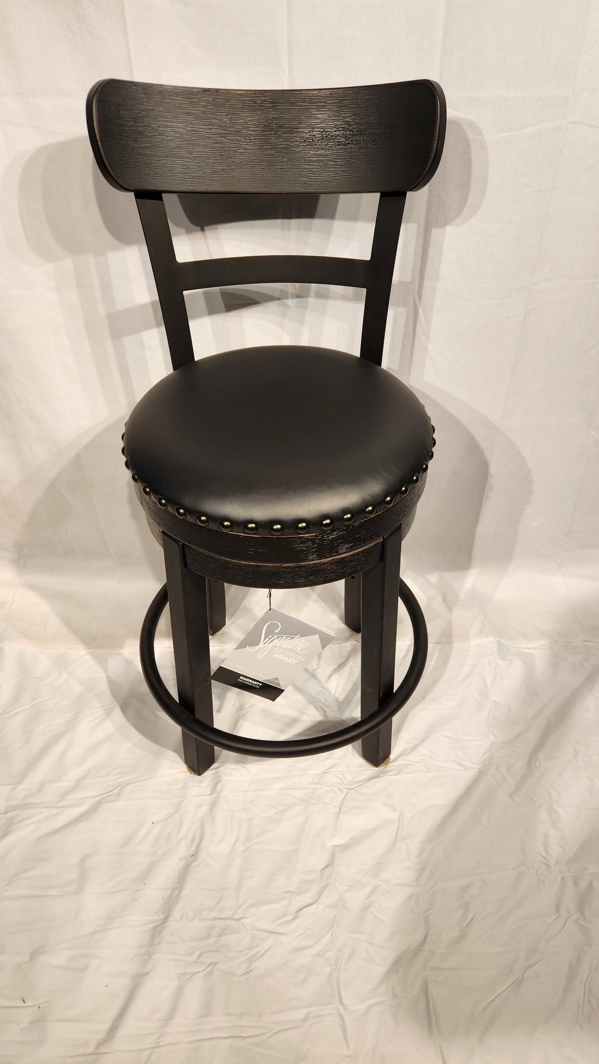 Ashley Valebeck D546: Black faux leather bar stool, round cushion seat, brass nailhead trim on white surface.