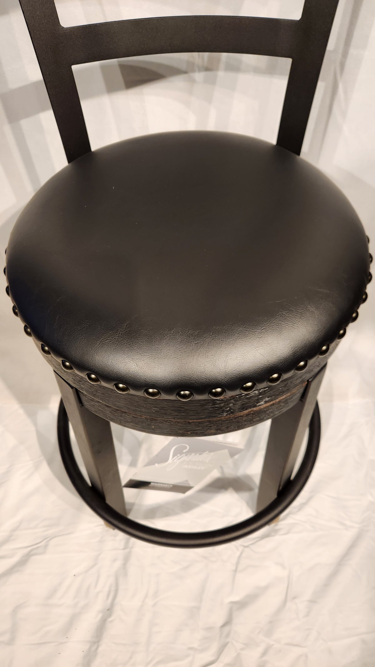 The Ashley Valebeck D546 black faux leather bar stool features a cushioned seat and metal studs, set on a white background.