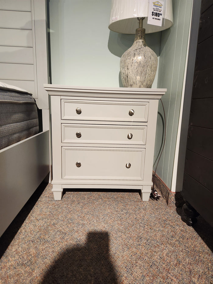 The Magnussen B5611 Charleston Nightstand, in white with three drawers, stands on carpet with a lamp on top.