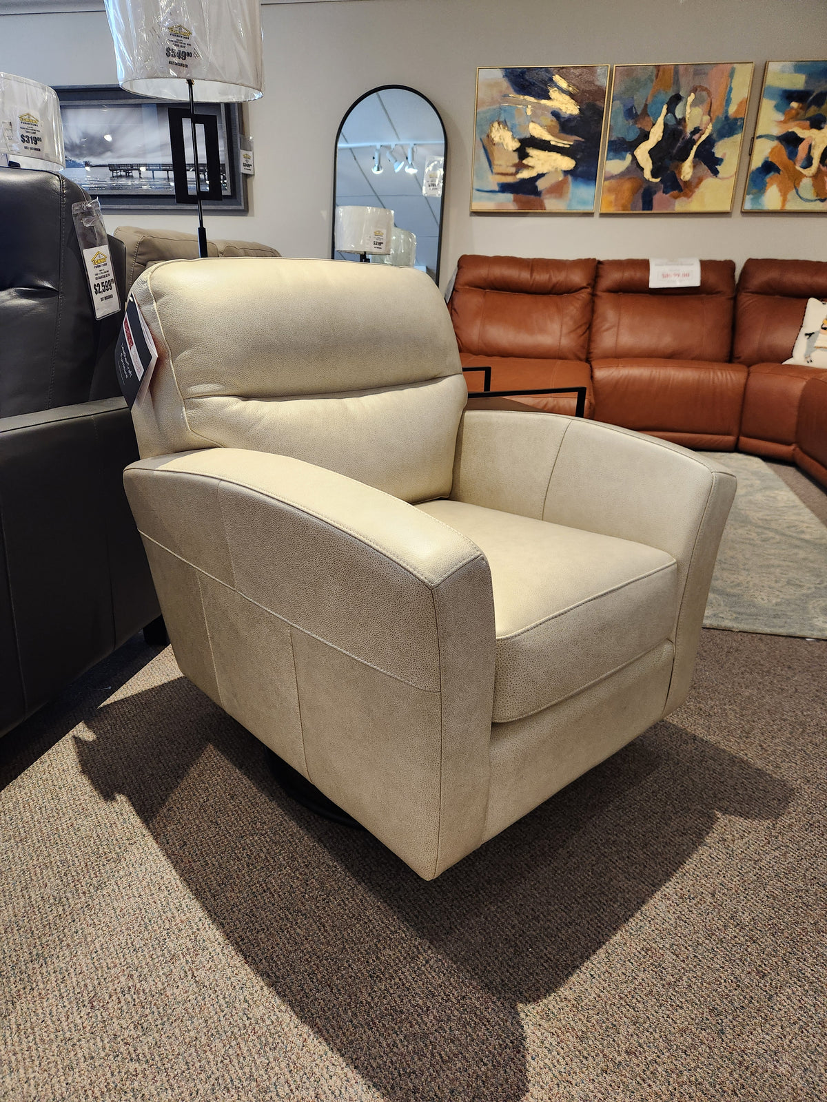 The Leather Craft 744 Leather Swivel Chair in beige is shown in a showroom with sofas and artwork behind it.