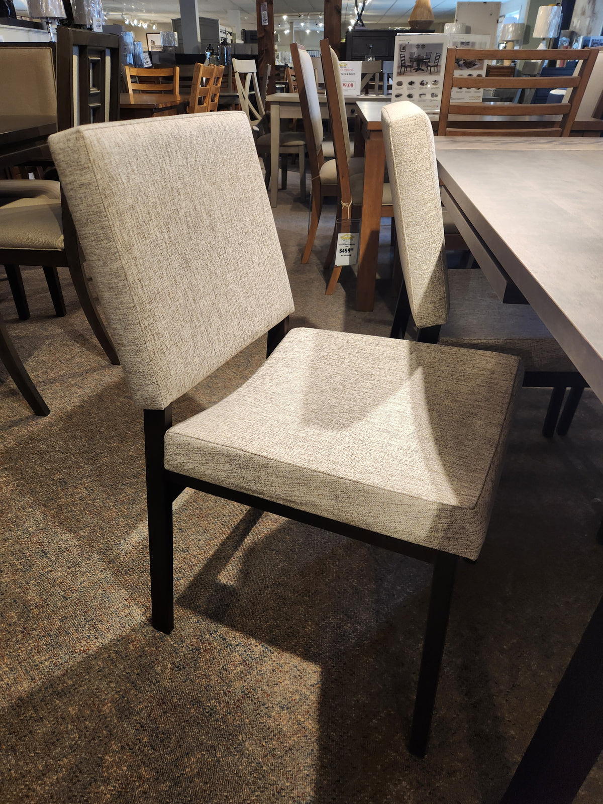 A beige dining chair with dark wooden legs beside the Amisco Lewis 36"X48" Table Package in a modern showroom.