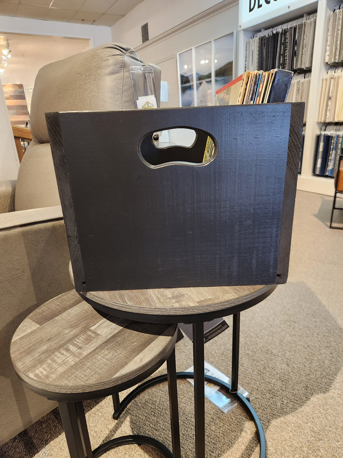 The SB101 Record Crate by Mercana is on a round side table near a gray armchair in a showroom with fabric displays.