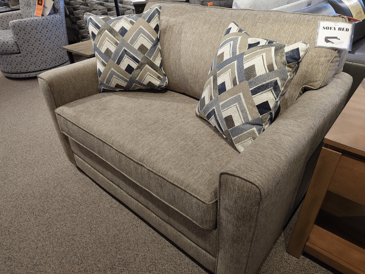 Simmons Upholstery 1014 Trinity Chair Bed in beige with two geometric pillows, shown in a furniture showroom.