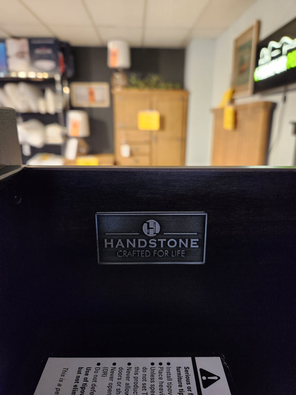 A close-up of a Handstone label on dark wood showcases the Stockholm 5 Drawer Lingerie Chest in a blurred showroom.