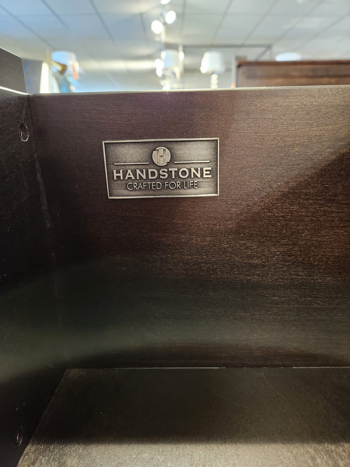 A metal "Handstone Crafted for Life" label accents the Stockholm 2-Drawer Nightstand’s sleek dark wood design.