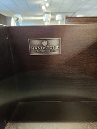 A metal "Handstone Crafted for Life" label accents the Stockholm 2-Drawer Nightstand’s sleek dark wood design.