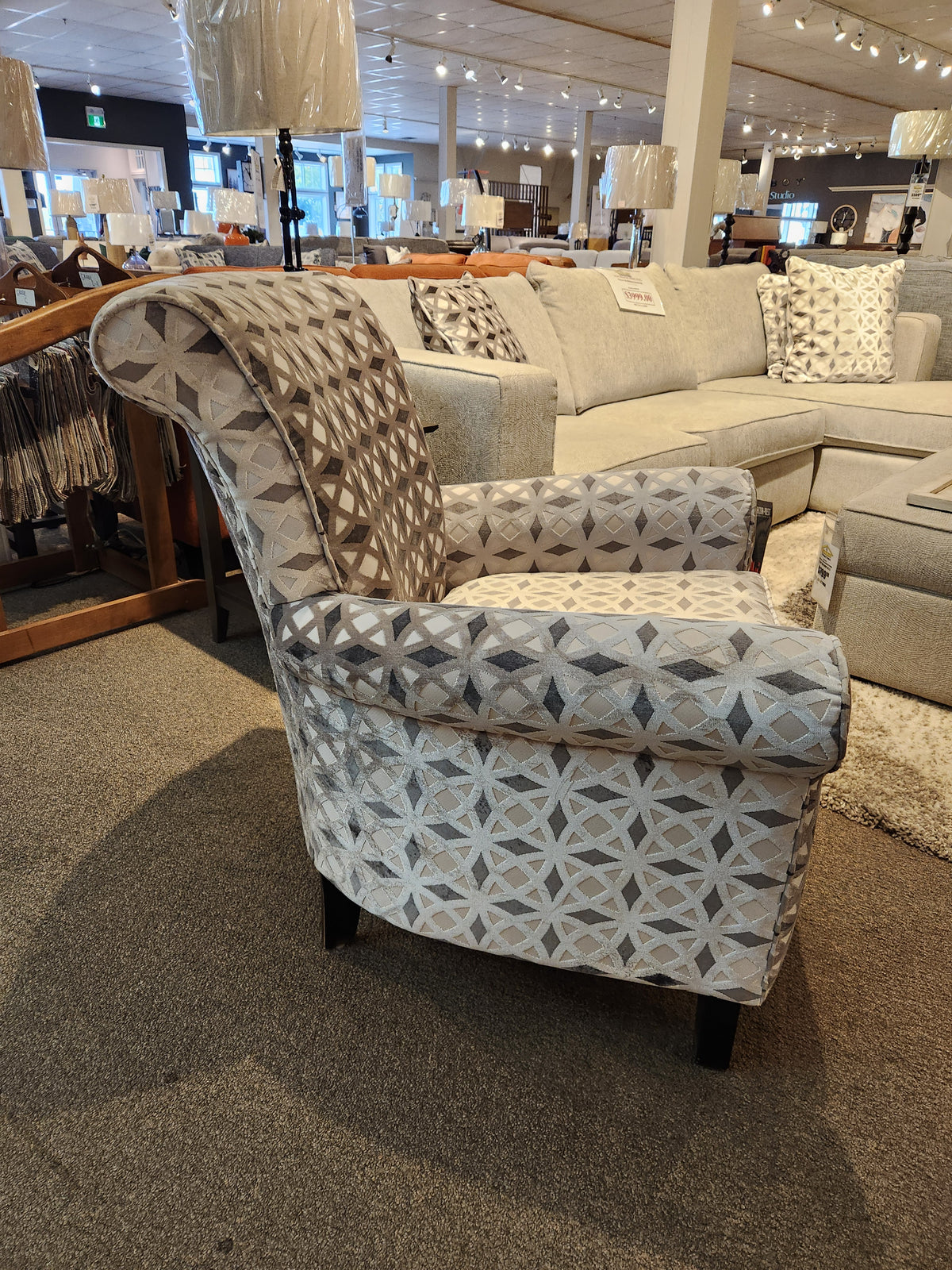 Explore the 2470 Stationary Accent Chair by Decor-rest, featuring elegant geometric patterned upholstery. Displayed in a showroom next to a gray sectional sofa and floor lamps, this Canadian-made accent chair effortlessly combines style and comfort, elevating the aesthetic charm of any living space.