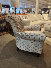 Explore the 2470 Stationary Accent Chair by Decor-rest, featuring elegant geometric patterned upholstery. Displayed in a showroom next to a gray sectional sofa and floor lamps, this Canadian-made accent chair effortlessly combines style and comfort, elevating the aesthetic charm of any living space.