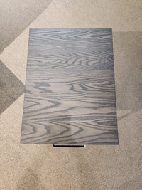 The Muskoka 18" End Table by Handstone features a solid oak top and gray wood grain, standing on a light carpeted floor.