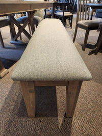 A beige bench with wooden legs sits on a carpet surrounded by the Handstone Retreat 7pc Dining Package.