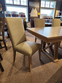 Gray upholstered chair near Handstone Retreat 7pc Dining Package in a showroom with artwork in the background.