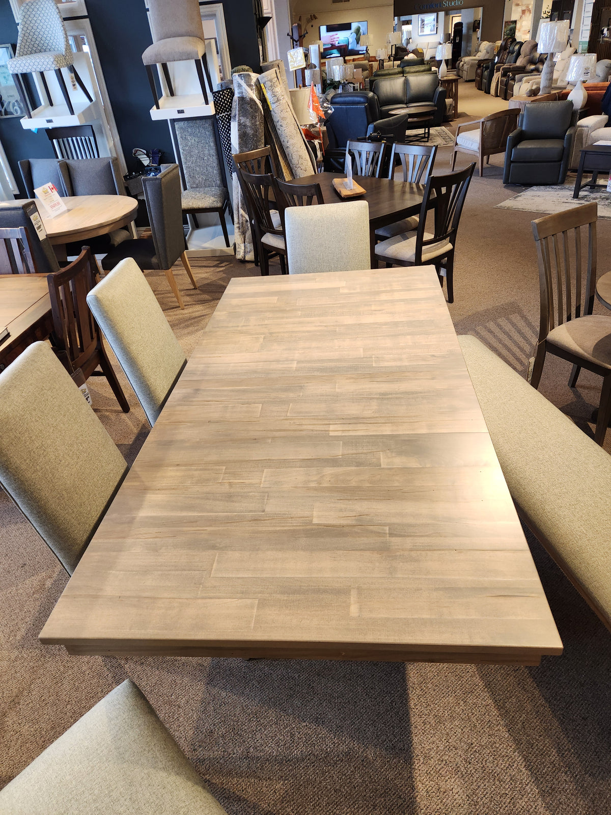 The Handstone Retreat 7pc Dining Package in a furniture showroom features a table and six upholstered chairs.
