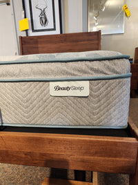 Close-up of a Beauty-rest Keegan Pillow-Top Plush Mattress with gray fabric and blue piping on a showroom bed frame.