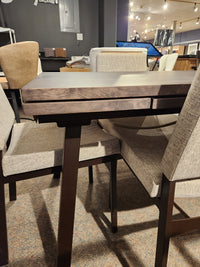 The Amisco Alex 36"x60" Solid Top Dining Package, featuring a dark wood top and four beige padded chairs, is on display.
